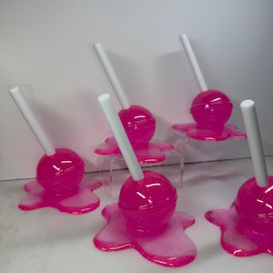 Puede incluir: Seis piruletas de color rosa brillante con palitos blancos. Cada piruleta se asienta sobre una base rosa brillante y derretida. Las piruletas est&aacute;n dispuestas sobre una superficie blanca, creando una exhibici&oacute;n l&uacute;dica y colorida.