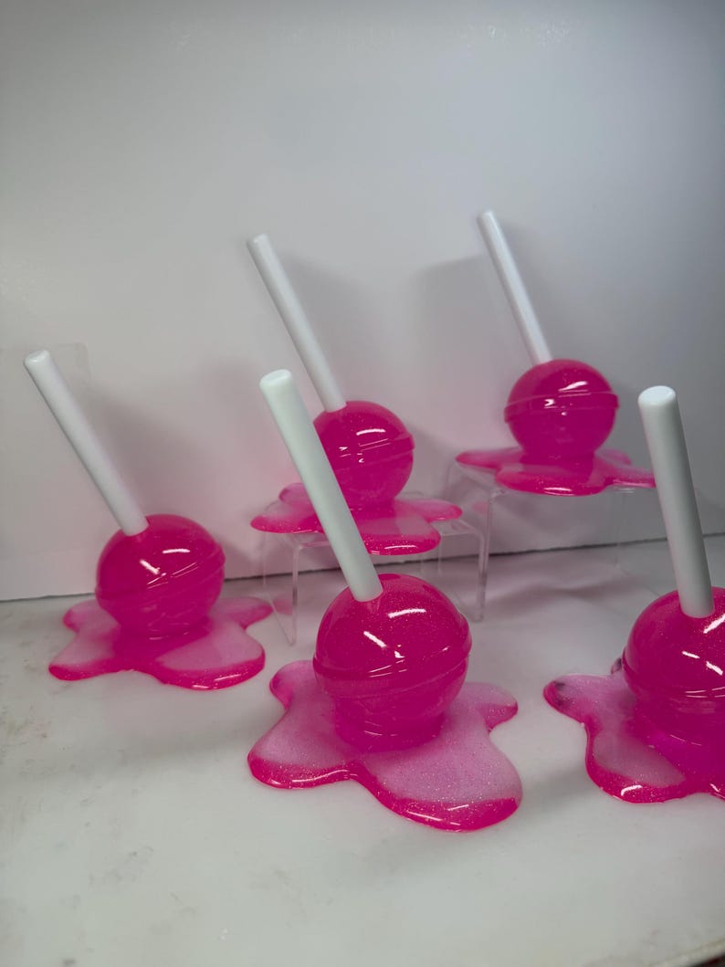 Puede incluir: Seis piruletas de color rosa brillante con palitos blancos. Cada piruleta se asienta sobre una base rosa brillante y derretida. Las piruletas est&aacute;n dispuestas sobre una superficie blanca, creando una exhibici&oacute;n l&uacute;dica y colorida.
