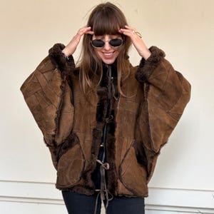 Puede incluir: Chaqueta de ante marrón con diseño de patchwork y ribete de piel sintética. La chaqueta tiene una silueta única tipo capa y presenta cierre de botón y detalle de lazo. El modelo lleva pantalones negros y zapatos negros.