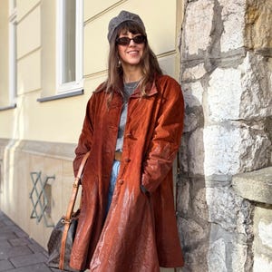 Może przedstawiać: Długi, rdzawy skórzany płaszcz z rozkloszowaną spódnicą i zapięciem na guziki. Płaszcz ma wygląd vintage i jest zestawiony z niebieskimi dżinsami i brązowymi butami. Brązowa skórzana torebka jest noszona na ramieniu.