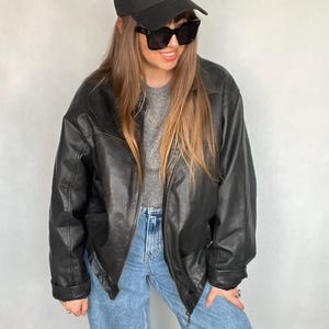 Puede incluir: Chaqueta de cuero negra con cremallera, combinada con vaqueros azul claro y una gorra de béisbol negra con letras blancas. El conjunto incluye gafas de sol negras y una camiseta gris.