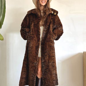 May include: A long, brown faux fur coat featuring a dark, repeating snakeskin pattern. The coat has a wide collar, long sleeves, and a straight-cut silhouette that extends to the mid-calf. It is worn over a white garment with dark brown boots.