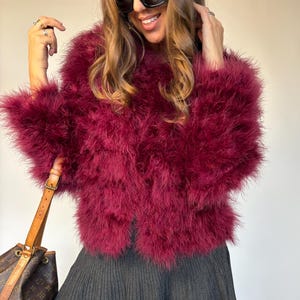 Könnte beinhalten: Burgunderfarbene Federjacke mit kurzem, ausgestelltem grauen Rock. Die Jacke hat eine flauschige Textur und einen runden Ausschnitt. Das Outfit wird mit schwarzen kniehohen Stiefeln und einer braunen Handtasche mit Logodruck kombiniert.