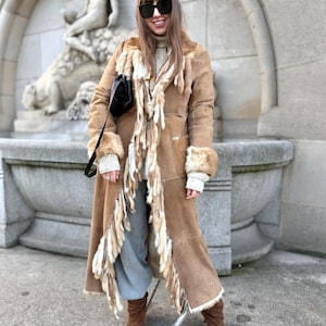 May include: A long, brown suede coat with fur trim and fringe detailing. The coat has a button-down front and is worn with brown boots. The coat's design includes fur cuffs and a decorative fringe. The coat is a classic style.