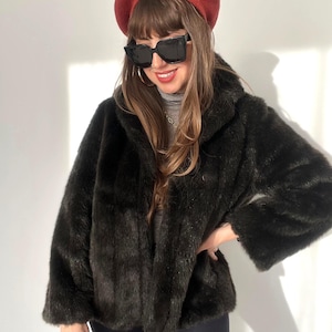 Puede incluir: Una chaqueta de piel sintética de color marrón oscuro con mangas largas y cuello ancho, combinada con una boina roja, gafas de sol cuadradas negras de gran tamaño, una blusa gris de cuello alto y un pequeño bolso de mano de color rojo.