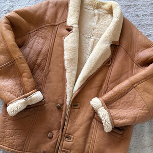 May include: A brown leather jacket with a cream-colored shearling lining and trim. The jacket features a button-up front, notched lapels, and shearling cuffs. The leather has a slightly distressed appearance.