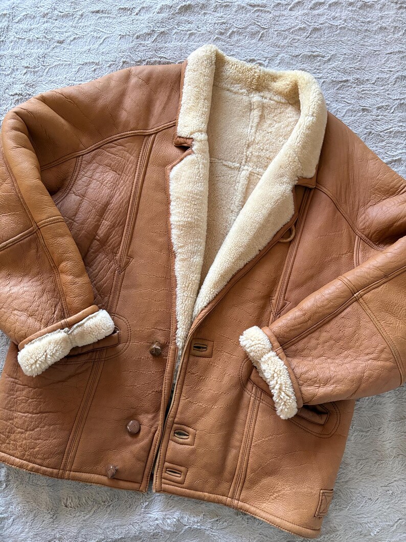 May include: A brown leather jacket with a cream-colored shearling lining and trim. The jacket features a button-up front, notched lapels, and shearling cuffs. The leather has a slightly distressed appearance.