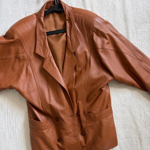 May include: A cognac-coloured leather blazer featuring a double-breasted front with four dark brown buttons, wide lapels, and two front pockets. The jacket has structured shoulders and a smooth, tan-coloured interior lining with a subtle diamond pattern.