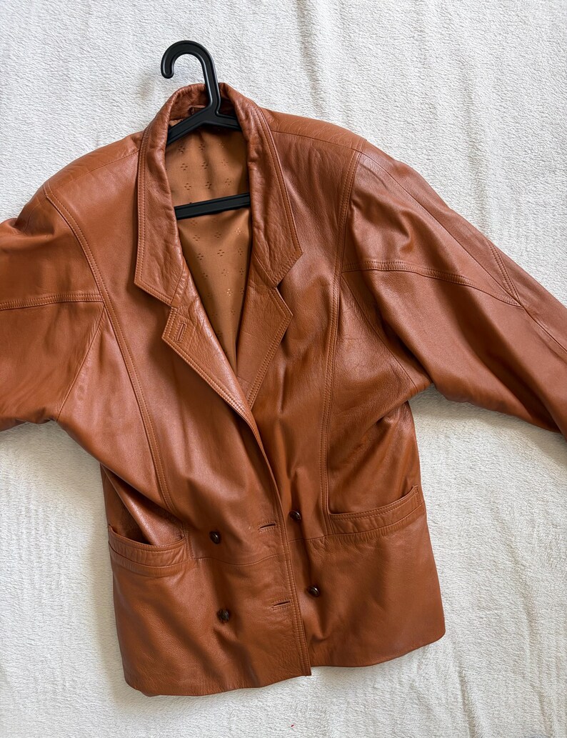 May include: A cognac-coloured leather blazer featuring a double-breasted front with four dark brown buttons, wide lapels, and two front pockets. The jacket has structured shoulders and a smooth, tan-coloured interior lining with a subtle diamond pattern.