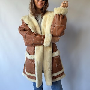 May include: A brown leather coat featuring a patchwork design with visible white stitching, a thick cream-colored shearling lining, a wide collar, and large cuffs. The coat includes two front pockets trimmed with shearling and blue denim jeans.