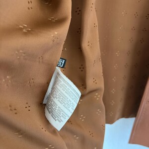 May include: A tan garment interior featuring a repeating pattern of small, perforated diamond shapes. A white care label with black text is attached to the inner seam, providing instructions in German regarding the care of lamb nappa leather.
