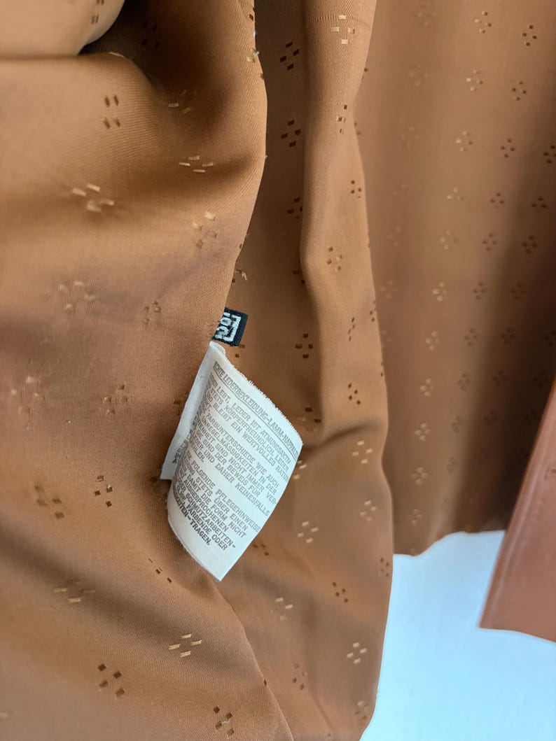 May include: A tan garment interior featuring a repeating pattern of small, perforated diamond shapes. A white care label with black text is attached to the inner seam, providing instructions in German regarding the care of lamb nappa leather.