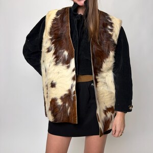 May include: A vintage cowhide vest with a brown and cream pattern. The open-front vest is worn over a black top and skirt. The vest's texture and color create a striking visual contrast.