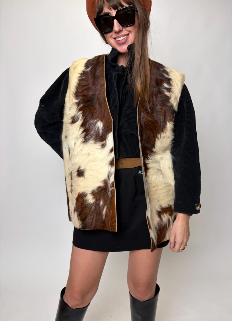 May include: A vintage cowhide vest with a brown and cream pattern. The open-front vest is worn over a black top and skirt. The vest's texture and color create a striking visual contrast.