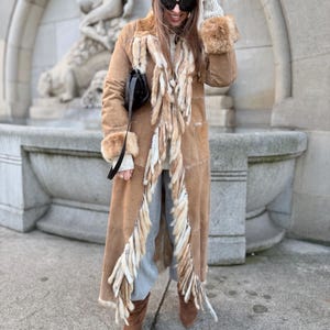 May include: A long tan suede coat featuring a prominent vertical trim of shaggy faux fur along the front opening and cuffs. The coat is worn over a light gray sweater and matching pants, paired with brown pointed-toe boots and a black shoulder bag.