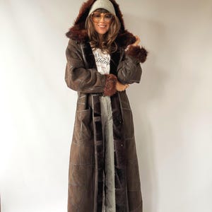 Può includere: Un lungo cappotto in camoscio marrone scuro con cappuccio bordato in pelliccia sintetica marrone scuro, polsini in pelliccia abbinati e cuciture a vista. Indossato sopra una t-shirt bianca con testo nero, pantaloni larghi chiari e stivali color cuoio.
