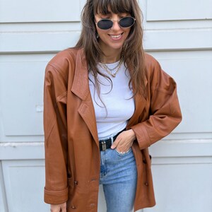 May include: A brown leather blazer with a double-breasted button closure, long sleeves, and notched lapels. The jacket is worn over a white ribbed top and light blue denim jeans with a black belt. Oval-shaped sunglasses are worn on the face.