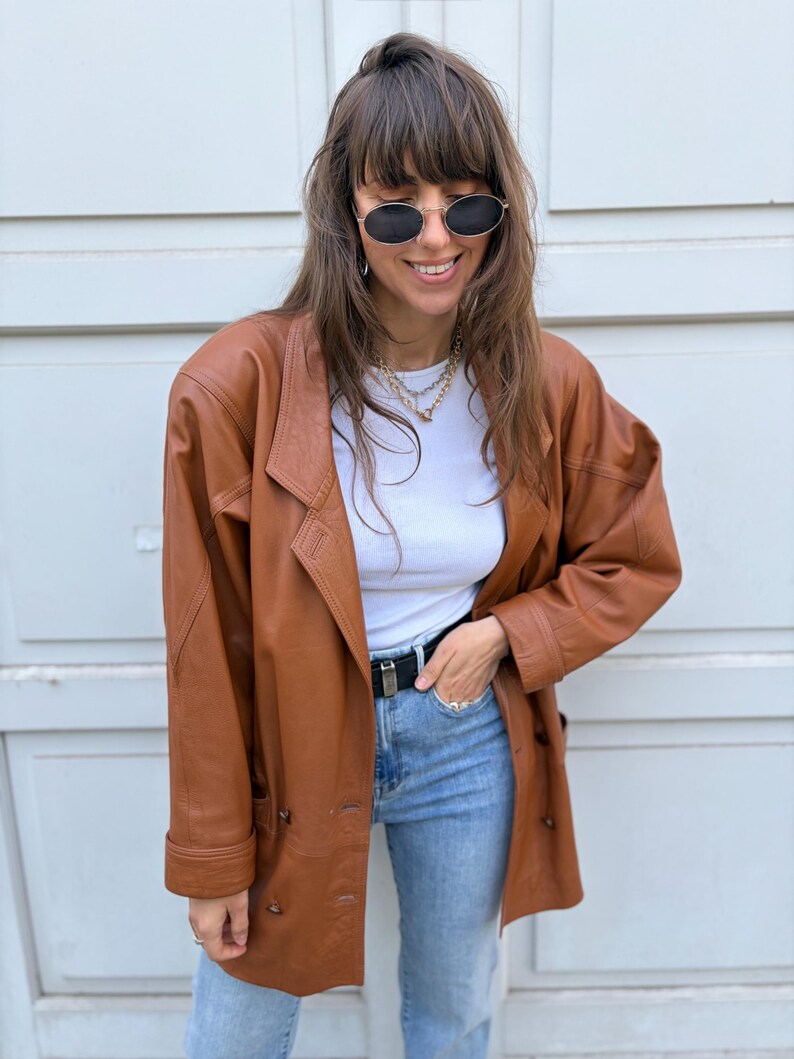 May include: A brown leather blazer with a double-breasted button closure, long sleeves, and notched lapels. The jacket is worn over a white ribbed top and light blue denim jeans with a black belt. Oval-shaped sunglasses are worn on the face.