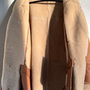 May include: A tan shearling coat with a brown leather exterior hangs on a black hanger. The coat's interior is a soft, light tan, contrasting with the darker brown leather trim and pockets. The coat is open, revealing the plush interior.