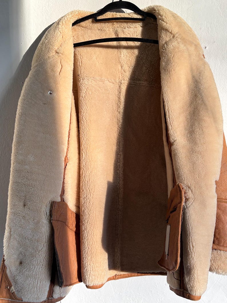 May include: A tan shearling coat with a brown leather exterior hangs on a black hanger. The coat's interior is a soft, light tan, contrasting with the darker brown leather trim and pockets. The coat is open, revealing the plush interior.
