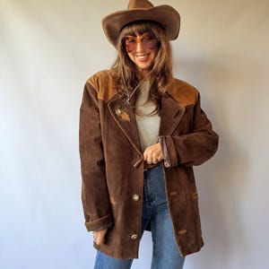 Pode incluir: Um casaco de camurça castanho com um jugo castanho mais escuro, gola de lapela e botões prateados. O casaco é usado sobre um top cinzento claro e calças de ganga azuis, combinado com um chapéu de cowboy de feltro castanho e óculos de sol âmbar.