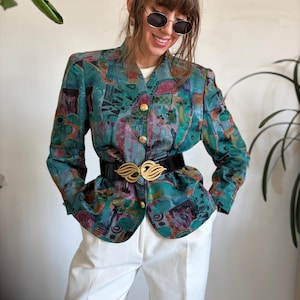 Könnte beinhalten: Ein Blazer mit teal-, lila- und grünen Mustern mit goldenen Knöpfen und einem schwarzen Gürtel mit goldener Schnalle. Der Blazer wird mit einer weißen Hose kombiniert. Das Outfit ist im Vintage-Stil.