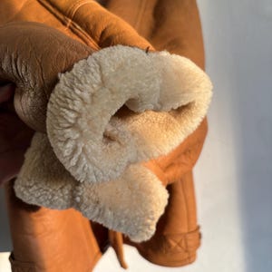 May include: Close-up of a brown leather jacket with a cream-colored, fluffy interior lining visible at the cuff. The jacket has a classic design with visible stitching and a textured leather exterior.