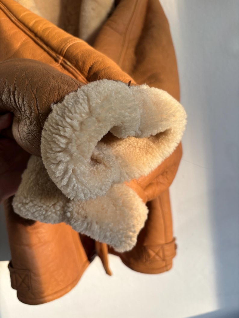 May include: Close-up of a brown leather jacket with a cream-colored, fluffy interior lining visible at the cuff. The jacket has a classic design with visible stitching and a textured leather exterior.