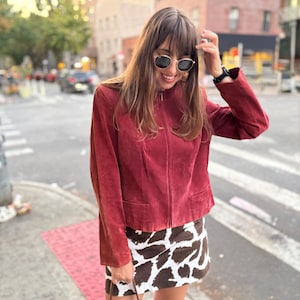 May include: A burgundy suede jacket with a zip-up front and two front pockets. The jacket is paired with a brown and white cow print mini skirt. The person is wearing sunglasses and a watch.
