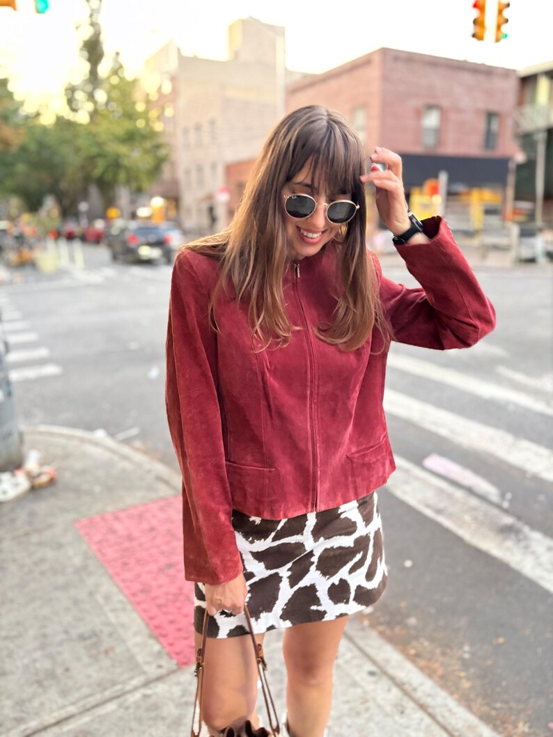 May include: A burgundy suede jacket with a zip-up front and two front pockets. The jacket is paired with a brown and white cow print mini skirt. The person is wearing sunglasses and a watch.
