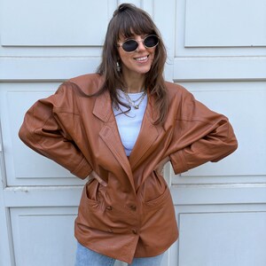 May include: A brown leather blazer featuring a double-breasted front with four buttons, wide lapels, and two front pockets. The jacket has long sleeves with stitched detailing at the cuffs and shoulders, paired with a white top and light blue jeans.