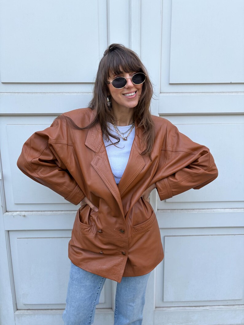 May include: A brown leather blazer featuring a double-breasted front with four buttons, wide lapels, and two front pockets. The jacket has long sleeves with stitched detailing at the cuffs and shoulders, paired with a white top and light blue jeans.