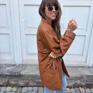 May include: A tan leather jacket with a collared neckline, long sleeves featuring wide cuffs, and two front pockets. The jacket has a smooth finish and a button-down front closure. The garment is paired with light blue denim jeans and dark sunglasses.