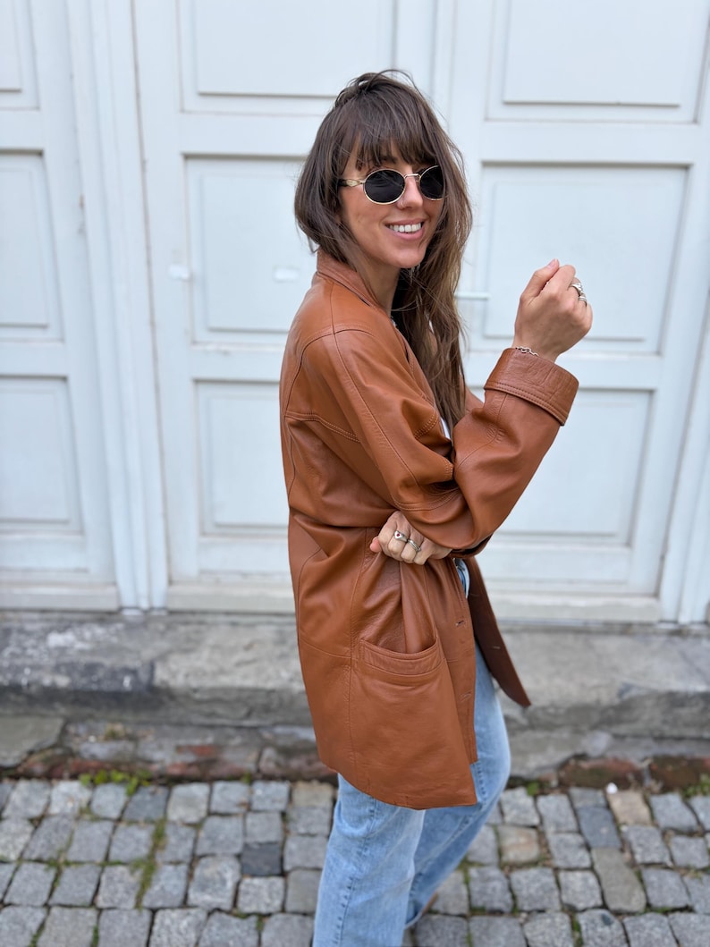 May include: A tan leather jacket with a collared neckline, long sleeves featuring wide cuffs, and two front pockets. The jacket has a smooth finish and a button-down front closure. The garment is paired with light blue denim jeans and dark sunglasses.