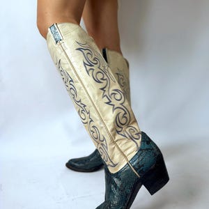 Könnte beinhalten: Kniehohe Westernstiefel mit einem cremefarbenen Lederschaft und aufwendigen, dunkelblau gestickten Flammenmustern. Der Fußbereich und die Ferse bestehen aus einem strukturierten, dunkel-türkisfarbenen Material in Schlangenlederoptik mit Blockabsatz.