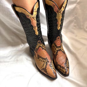 Puede incluir: Un par de botas de vaquero negras con un estampado de piel de serpiente en la puntera y la parte superior de la ca&ntilde;a. Las botas tienen una puntera puntiaguda y costuras decorativas. El estampado de piel de serpiente presenta tonos marrones, amarillos y rosas.
