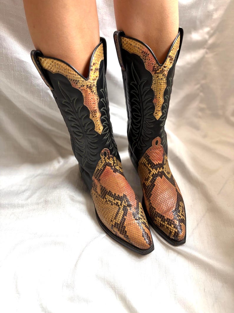 Puede incluir: Un par de botas de vaquero negras con un estampado de piel de serpiente en la puntera y la parte superior de la ca&ntilde;a. Las botas tienen una puntera puntiaguda y costuras decorativas. El estampado de piel de serpiente presenta tonos marrones, amarillos y rosas.