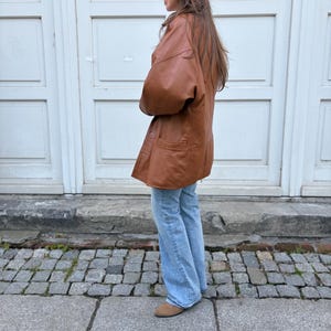 May include: A tan leather jacket with a button-front closure and two front pockets, paired with light blue denim jeans and tan suede shoes. The jacket features long sleeves and a relaxed fit, worn over straight-leg jeans that reach the ankles.