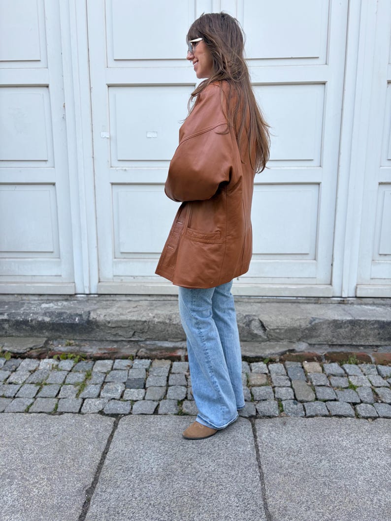 May include: A tan leather jacket with a button-front closure and two front pockets, paired with light blue denim jeans and tan suede shoes. The jacket features long sleeves and a relaxed fit, worn over straight-leg jeans that reach the ankles.