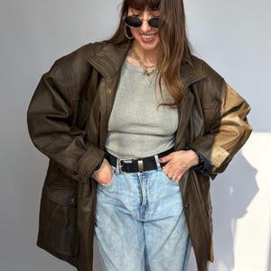 K&ouml;nnte beinhalten: Braune Lederjacke mit klassischem Design. Die Jacke hat einen Kragen, Kn&ouml;pfe und mehrere Taschen. Das Model tr&auml;gt ein graues Oberteil und eine hellblaue Jeans. Ein schwarzer G&uuml;rtel und eine Sonnenbrille vervollst&auml;ndigen den Look.