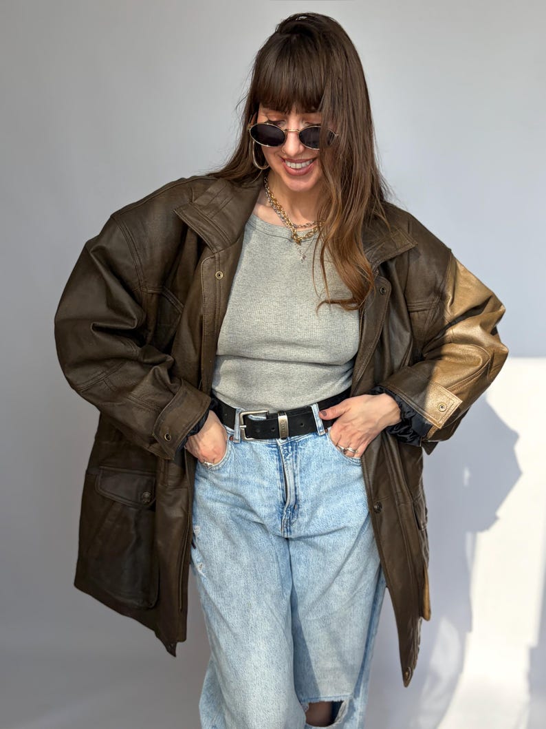 K&ouml;nnte beinhalten: Braune Lederjacke mit klassischem Design. Die Jacke hat einen Kragen, Kn&ouml;pfe und mehrere Taschen. Das Model tr&auml;gt ein graues Oberteil und eine hellblaue Jeans. Ein schwarzer G&uuml;rtel und eine Sonnenbrille vervollst&auml;ndigen den Look.