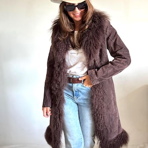 Può includere: Un cappotto lungo in camoscio marrone scuro con un bordo in pelliccia folta e arruffata lungo il colletto, l'apertura frontale e l'orlo. Il cappotto presenta un motivo geometrico cucito sul corpo e sulle maniche, abbinato a jeans azzurri.