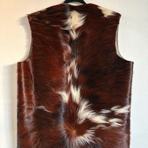 May include: A sleeveless vest made of cowhide. The vest features a brown and white pattern, with the brown dominating the color scheme. The vest is hanging on a black hanger against a white wall. The back of the vest is visible.