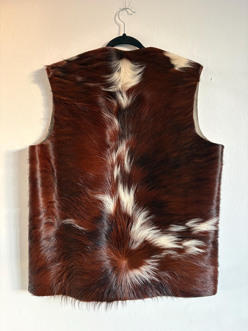 May include: A sleeveless vest made of cowhide. The vest features a brown and white pattern, with the brown dominating the color scheme. The vest is hanging on a black hanger against a white wall. The back of the vest is visible.