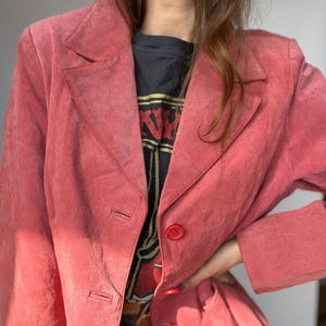 May include: A dusty rose blazer with a classic notched lapel and two-button closure. The jacket has long sleeves and a tailored fit. The blazer is made of a soft, suede-like material. The blazer is a vintage piece.