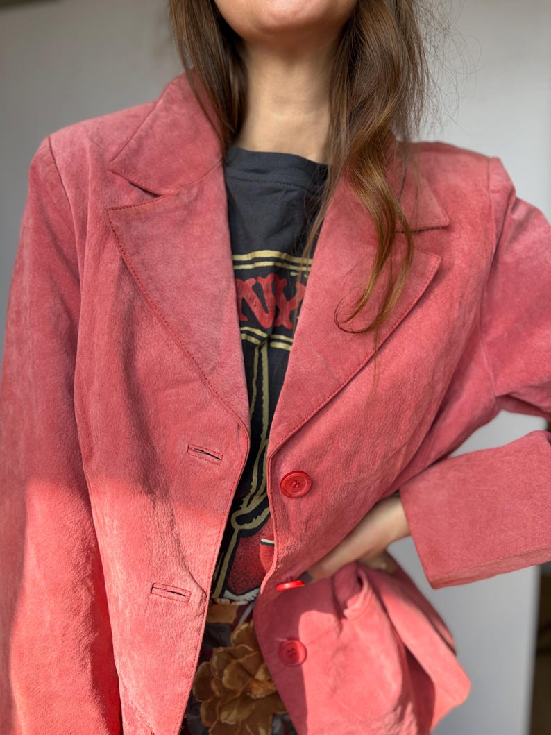 May include: A dusty rose blazer with a classic notched lapel and two-button closure. The jacket has long sleeves and a tailored fit. The blazer is made of a soft, suede-like material. The blazer is a vintage piece.