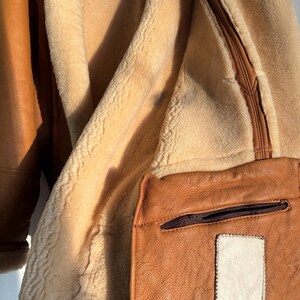 May include: Close-up of a brown leather jacket with a cream-colored shearling lining. The jacket features a small, square pocket with a zipper and a white patch. The leather has a textured appearance.