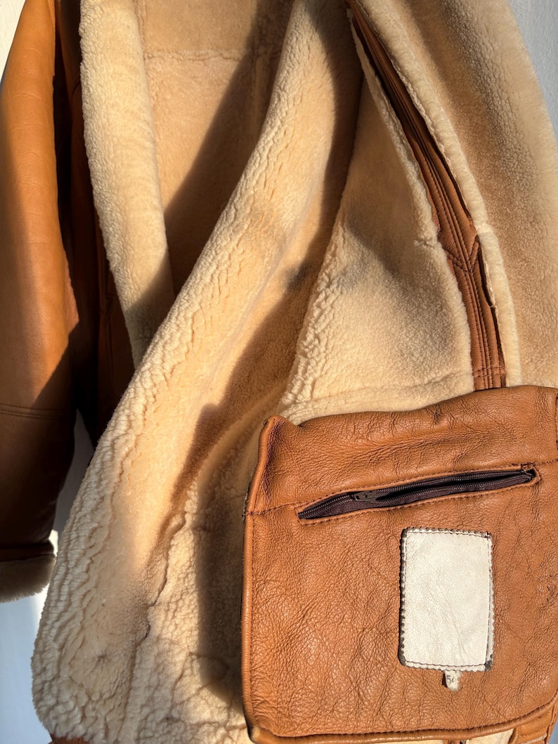 May include: Close-up of a brown leather jacket with a cream-colored shearling lining. The jacket features a small, square pocket with a zipper and a white patch. The leather has a textured appearance.