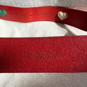 May include: A red leather belt with heart-shaped embellishments. One heart is green, and the other is cream-colored. The belt is marked "MADE IN ITALY" and "VERA PELLE". The size is 80.