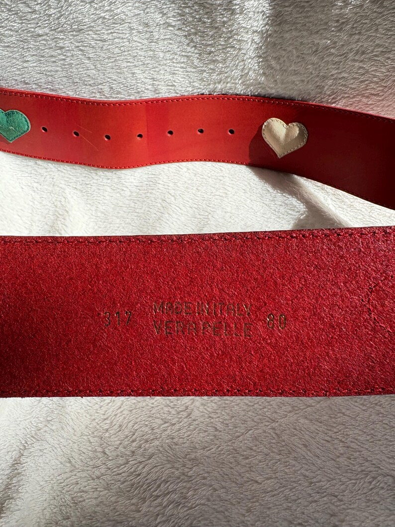 May include: A red leather belt with heart-shaped embellishments. One heart is green, and the other is cream-colored. The belt is marked "MADE IN ITALY" and "VERA PELLE". The size is 80.